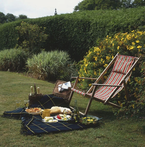 Striped deck chair on grass by picnic blanket spread with plates of food and bottles of wine, side view