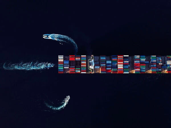 Aerial view of a cargo ship loaded with containers being instructed by two pilot boats