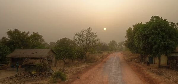 Local road in rural Gambia with farmhouse, forest and sunrise sun on hazy morning