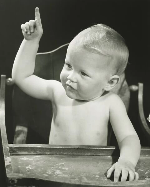 Baby boy (6-12 months) pointing, sitting in high chair, (B&W), portrait