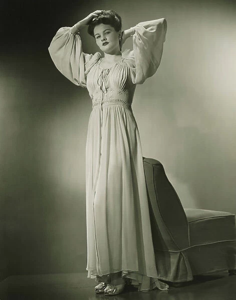 Woman in nightdress standing by chair in studio, (B&W)