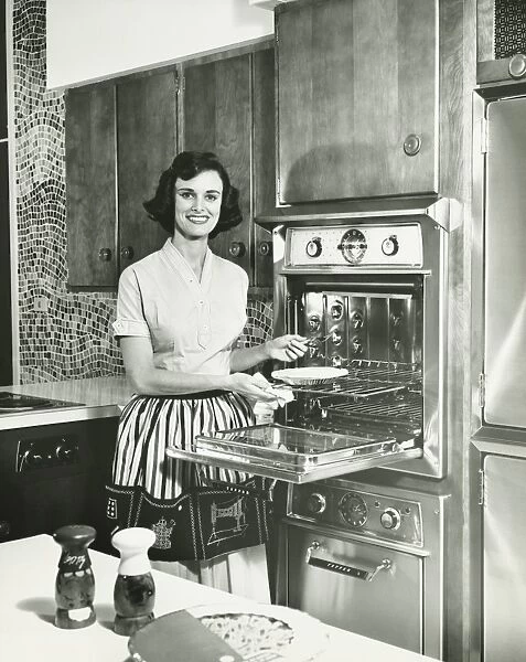 Woman putting pie into oven in kitchen, (B&W), portrait