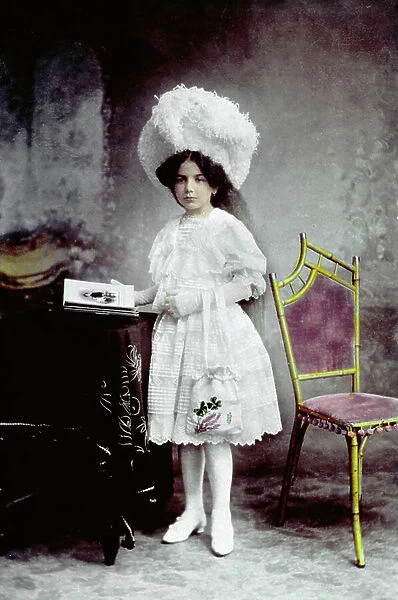 Full-length hand-colored portrait of little girl. She is standing beside a chair and a table on which there is a photograph album. She is wearing an elegant white dress and an opulent plumed hat