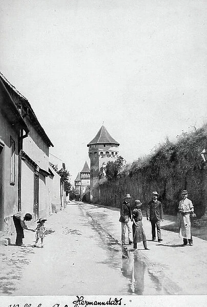 A group of people resting on a street in Sibiu (formerly Hermannstadt) in Romania. On the background fortified towers in the city walls are visible