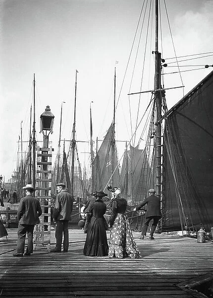 Spectators at the end of the wooden dock, looking at a forest of masts and sails in the port of Lowestoft (Suffolk, England), next to onlookers, two men pull a mooring rope, 1908 (b / w photo)
