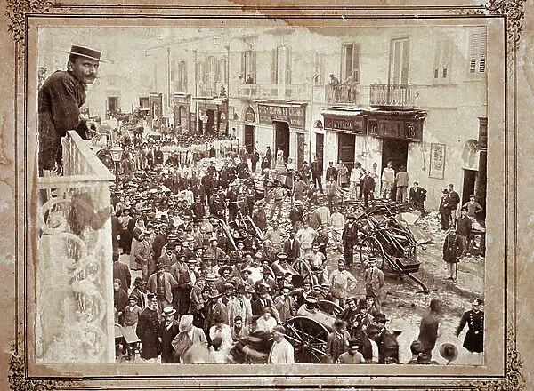 Street scene in an Italian city, a man on a balcony observes the crowd in the street, among which is found firefighters and law enforcement forces. Early 20th century