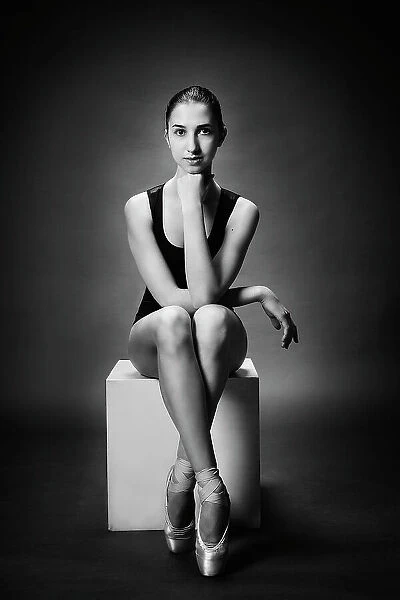 ballerina. A ballerina in a bodysuit and pointe shoes sits on a cube and looking directly