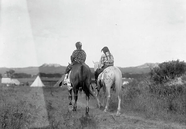 Two daughters of a chief on horseback, riding away from camera toward tents in background, c1907. Creator: Edward Sheriff Curtis