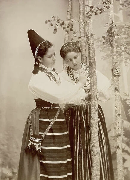 Two young women pose in different national costumes, 1890-1920. Creator: Helene Edlund