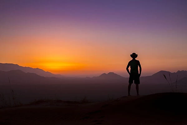 Silhouette of man wearing a cowboy hat looks out at the glowing landscape and sky at sunset, Namibia