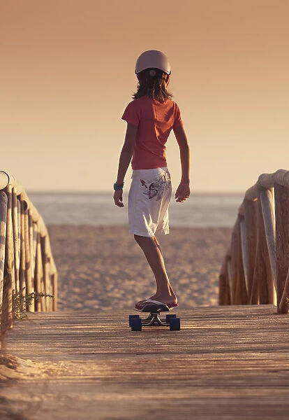 A Young Person Skateboarding With Bare Feet Over A Wooden Boardwalk Towards The Beach; Tarifa, Cadiz, Andalusia, Spain