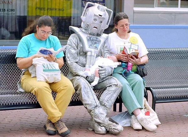 Person in a robot outfit sits down on a bench next to two women shoppers reading their