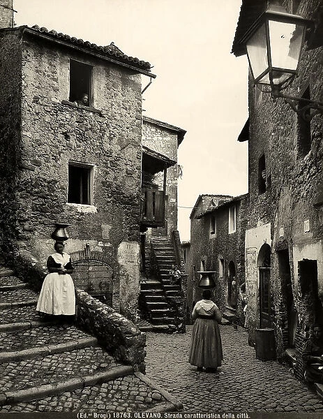 Busy street of Olevano Romano; on the stairs of the houses, one can see some inhabitants of the town and two women in common clothing with amphoras on their heads