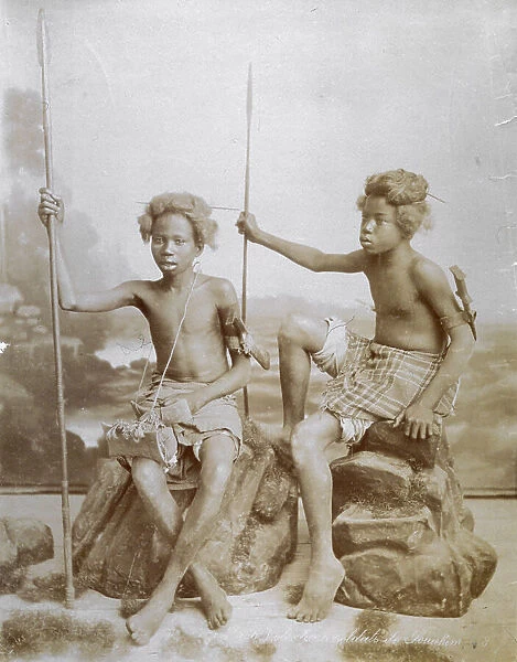 Full-length portrait of two african adolescents. They are bare from the waist up. Tied to the left arm is a dagger and in their right hand each one holds a lance. They are seated on papier-mache rocks