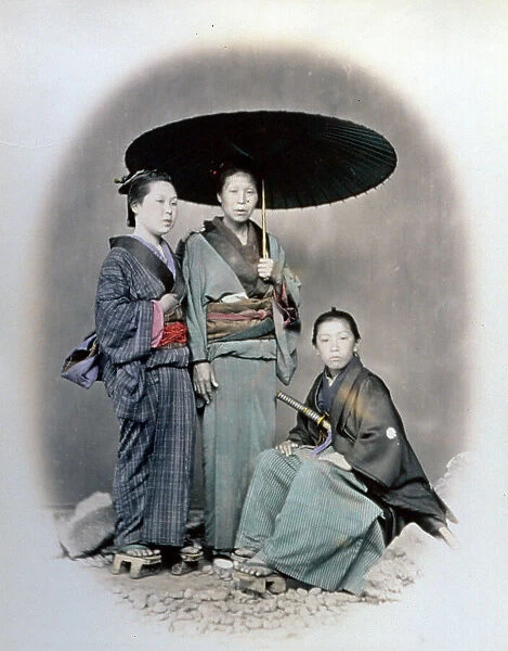 Full-length portrait of a japanese woman surrounded by her children. The mother and the young daughter wear the traditional kimono. The mother, at the center of the portrait, has an open umbrella in her right hand. The son in samurai dress is sitting on a rock