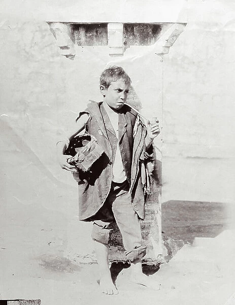 Full-length portrait of a neapolitan urchin in dirty ragged clothing. The sulky boy is holding a pipe in his left hand and has a basket under his right arm