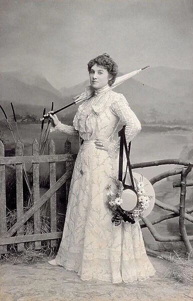 Full-length portrait of young woman elegantly dressed. The young woman is leaning on a wooden fence, behind her the backdrop of a landscape with a lake and mountains. In her left hand she has a closed umbrella resting on her shoulder, a straw hat decorated with daisies is tied to her right arm