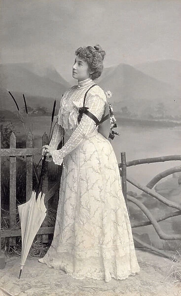 Full-length studio portrait of a young woman in three quarter view. She is leaning on a closed umbrella and is standing beside a stockade beyond which a backdrop can be observed of a landscape with a lake and mountains