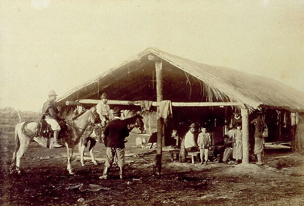 Group of persons in a hut with a straw roof, in Argentina. Outside two men on horseback dressed as gauchos and a third man seen from the back
