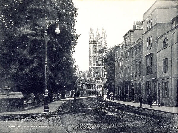 High Street full of pedestrians, in Oxford. The street is lined on the right by buildings, on the left by trees and a park. Beyond these, in the background, the 'Bell Tower', the tower on the facade of Magdalen College. In the foreground clearly visible on the left is a street lamp