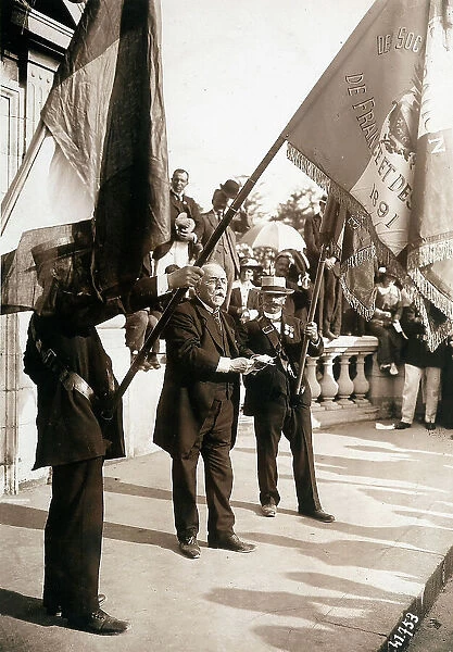 The image shows the Alsatians - Lorrainers at the statue of Strasbourg, in Paris. W. Saimboef makes a speech between two men who raise a flag. The episode occurs on 10 August 1914