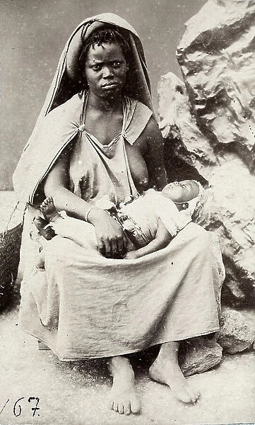 The image shows a young lady feeding her child. The lady, inhabitant of Khrumiria, Tunisia, wears a dress open over the breasts and a veiled head gear. Behind, a rocky setting