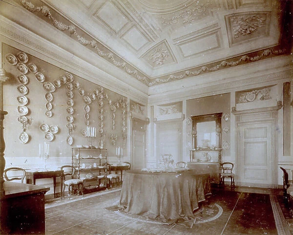 Interior of an elegant Nineteenth century house. At the center of the room is a table covered with a tablecloth. At the sides furniture and chairs. On the walls decorated plates are arranged in a pattern