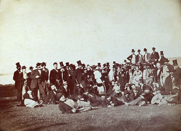 Large group of ladies and elegant gentlemen shown in a field. Some of the men are reclining on the grass in a relaxed position. On the right a man is playing a guitar