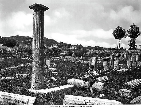 The remains of the Gymnasium, Olympia. On the right is column, almost intact, that made up part of the portico. Next to it is a youth sitting, playing what looks like a flute