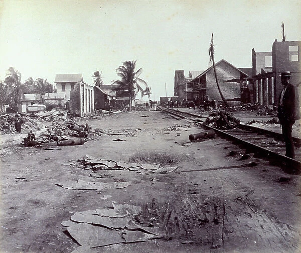 A street in Colon (Panama), strewn with rubble. In the foreground railroad tracks and on the right a man in a straw hat. In the background a few houses