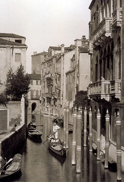 A venetian calle with a gondola going by. Other vessels are drawn up along the buildings. In the foreground a few mooring poles
