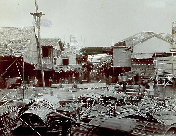 View of a corner of Canton near the river. In the foreground many wooden boats are moored and delapidated buildings. Men and children add life to the scene
