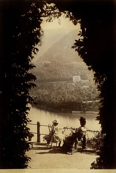 View of the lake of Como framed by the plants of the garden of Villa Serbelloni in Bellagio; near the balustrade a little girl and a lady are sitting