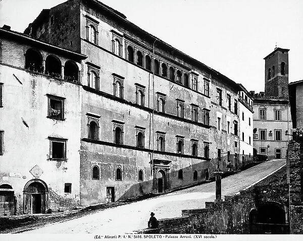 View of Palazzo Arroni in Spoleto. The building is decorated with an elegant entrance, two rows of windows and a loggia above. The facade is adorned with numerous graffiti. In the foreground is a man seated on a wall that runs along the street in front of the palazzo