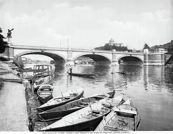 View of the River Po with some rowers and a boatman passing underneath the Umberto I bridge. On the dry gravel river-bed are some rowboats. In the background the Mount Cappuccini is visible with the Church of S. Maria del Monte