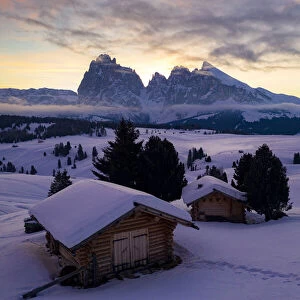 Wood cabins covered with snow with Sassopiatto and Sassolungo in background at dawn
