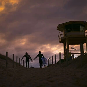 A pair of female surfers march up the beachhead alongside a lifesavers hut at sunset