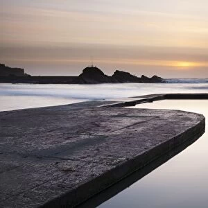 View of tidal swimming pool with incoming tide and waves breaking over breakwater at sunset, Bude Sea Pool