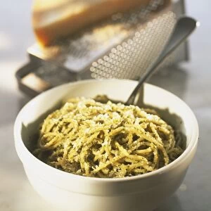 Bowl of spaghetti in pesto sauce sprinkled with grated cheese, block of cheese on grater in the background, close up