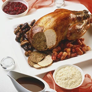 Christmas roast turkey served with rolled streaky bacon rashers and chipolatas, section of turkey sliced away revealing stuffing and cooked turkey meat. Jug of gravy, bread sauce and cranberry sauce served separately