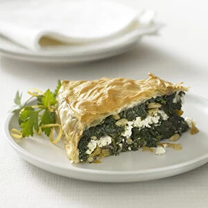 Filo pastry pie stuffed with spinach, feta cheese and pine nuts and garnished with parsley and lemon zest, plates and napkin in background, close-up