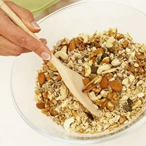 Hand using wooden spoon to stir bowl of muesli containing oats, brazil nuts, almonds, cashew nuts, walnuts and seeds