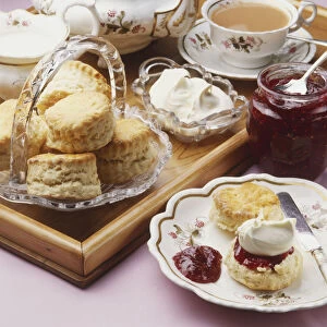 Scones, tea and clotted cream on a serving tray, and, next to it, a jar of jam and a scone topped with jam and cream on a plate