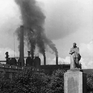 A statue of joseph stalin in the garden of victory square in the industrial city of stalinsk, the iron and steel plant is in the background, spewing smoke, early 1950s
