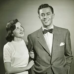Young couple arm in arm in studio, woman looking at man, smiling, (B&W)