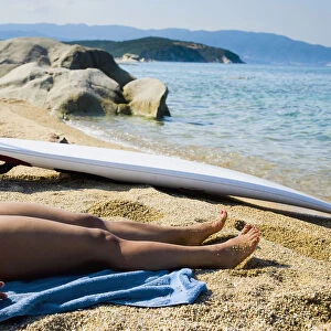 Greece, Halkidiki, Young woman sunbathing on beach with surf board on sand; Ierissos