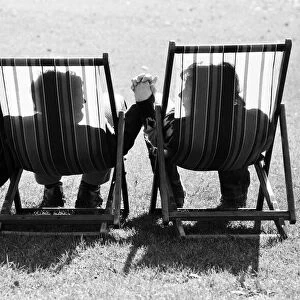 A couple in sit in a pair of deckchairs holding hands and looking at one another as they