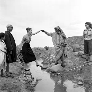 Man in arabic dress lends his hand to help French actresses across a stream as he guides