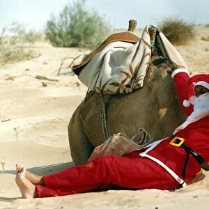 Santa Claus cools off by sitting in the shade of a camel in the desert on his way to