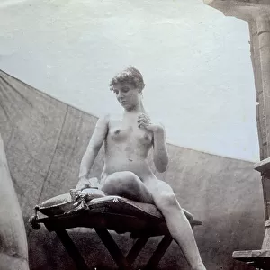 Portrait of a young nude woman. The model is posing in a statuesque pose, seated on a table covered with cushions and her face slightly inclined towards her breast and with her left hand on her breast. A curtain acts as background